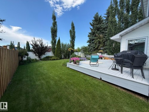 Expansive backyard featuring a manicured lawn, a large wood deck, mature trees, a wooden privacy fence, and a white stucco wall - 5604 57 Avenue, Beaumont, AB 