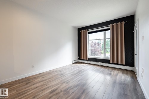 Spacious room featuring wood-finish flooring, white walls, and a prominent window with a patterned accent wall - #215 2590 Anderson Wy Sw, Edmonton, AB - Indoor Photo Showing Other Room