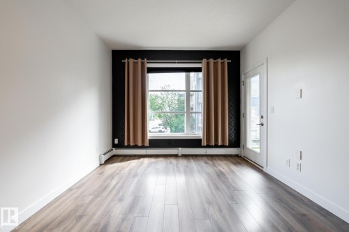 Spacious room featuring wood-finish flooring, a large window with exterior scenery, a glass-paneled door, baseboard heating, and a textured accent wall - #215 2590 Anderson Wy Sw, Edmonton, AB - Indoor Photo Showing Other Room