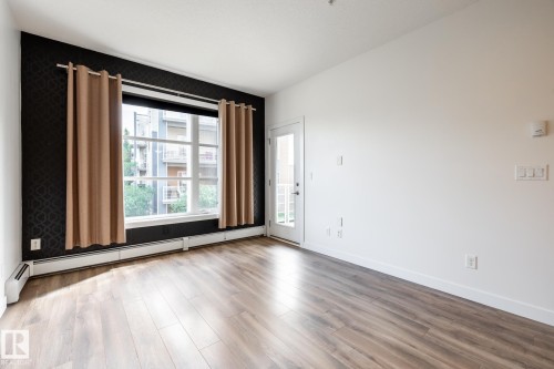 Spacious room with wood-finish flooring, a large window with baseboard heating, a white door leading to an exterior space, and a distinct patterned accent wall - #215 2590 Anderson Wy Sw, Edmonton, AB - Indoor Photo Showing Other Room