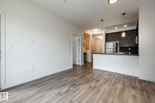 Open-concept living space featuring wood-finish flooring, a kitchen island with pendant lighting, dark wood-tone cabinetry, a stainless steel refrigerator, and a black subway tile backsplash - #215 2590 Anderson Wy Sw, Edmonton, AB - Indoor Photo Showing Kitchen