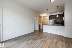 Open-concept living space featuring wood-finish flooring, a kitchen island with pendant lighting, dark wood-tone cabinetry, a stainless steel refrigerator, and a black subway tile backsplash - 