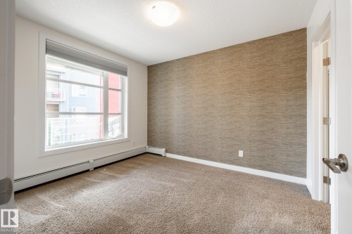 Neutral-toned wall covering, broad window, baseboard heating, recessed ceiling fixture, and textured carpeting - #215 2590 Anderson Wy Sw, Edmonton, AB - Indoor Photo Showing Other Room