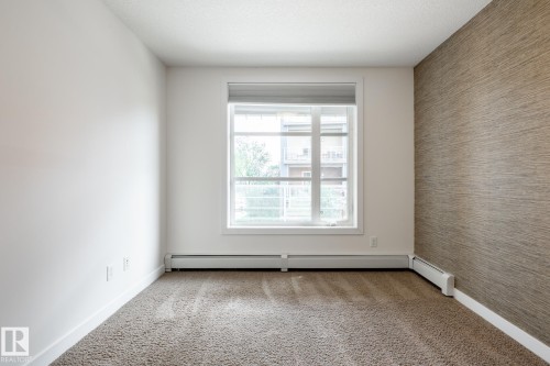 Bright room featuring a large window with white trim, tan textured accent wall, light-colored carpeting, and white baseboard heaters - #215 2590 Anderson Wy Sw, Edmonton, AB - Indoor Photo Showing Other Room