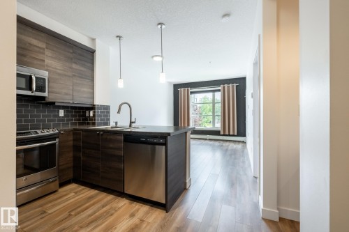 Modern kitchen featuring stainless steel appliances, dark wood-finish cabinetry, a subway tile backsplash, and a large island with an undermount sink - #215 2590 Anderson Wy Sw, Edmonton, AB - Indoor Photo Showing Kitchen With Upgraded Kitchen