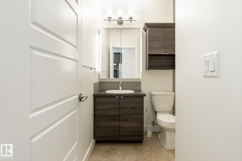 Bathroom featuring a wood-finish vanity with an integrated sink, a rectangular mirror with backlighting, a three-light vanity fixture, and a wall-mounted storage cabinet - #215 2590 Anderson Wy Sw, Edmonton, AB - Indoor Photo Showing Bathroom