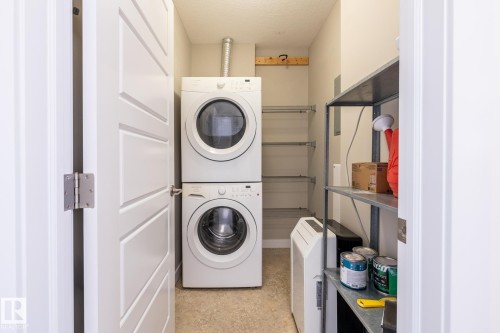 Dedicated laundry area featuring a stacked washer and dryer, metal shelving, neutral wall color, and textured flooring - #215 2590 Anderson Wy Sw, Edmonton, AB - Indoor Photo Showing Laundry Room