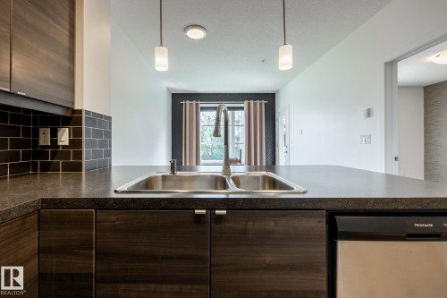 Kitchen featuring a dual-basin stainless steel sink with a high-arc faucet, dark wood-finish cabinetry, a black subway tile backsplash, and dark countertops - #215 2590 Anderson Wy Sw, Edmonton, AB - Indoor Photo Showing Kitchen With Double Sink