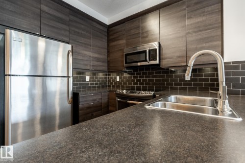 Kitchen featuring dark wood-finish cabinetry, stainless steel appliances, black subway tile backsplash, a dual basin stainless steel sink, and dark flecked countertops - #215 2590 Anderson Wy Sw, Edmonton, AB - Indoor Photo Showing Kitchen With Double Sink With Upgraded Kitchen