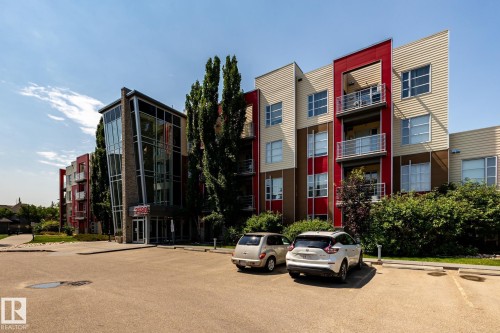Contemporary facade featuring a prominent glass entrance tower, red and beige paneling, multiple balconies with metal railings, and an expansive asphalt parking area - #215 2590 Anderson Wy Sw, Edmonton, AB - Outdoor With Facade