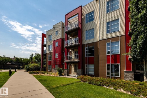 Multi-story building featuring modern red, tan, and brown exterior panels, large windows, and multiple private balconies with metal railings - #215 2590 Anderson Wy Sw, Edmonton, AB - Outdoor With Facade