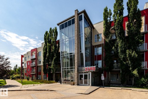 Contemporary multi-story building featuring a prominent glass entrance tower, red and beige exterior siding, multiple private balconies, a paved drive, and a manicured lawn - #215 2590 Anderson Wy Sw, Edmonton, AB - Outdoor With Facade