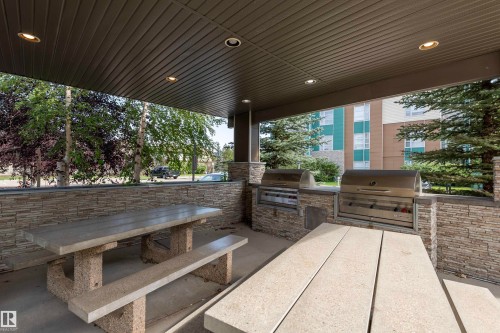 Covered outdoor entertaining area featuring two built-in stainless steel grills, stone-clad grill surrounds, recessed ceiling lighting, and multiple concrete picnic tables - #215 2590 Anderson Wy Sw, Edmonton, AB - Outdoor With Deck Patio Veranda