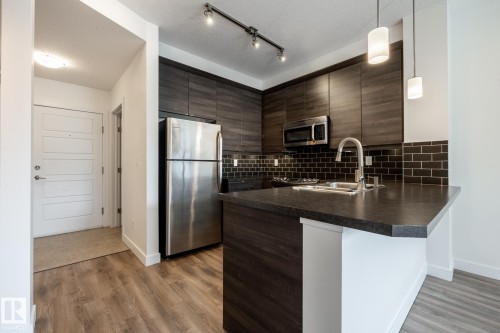 Kitchen featuring dark wood-finish cabinetry, stainless steel appliances, a dark countertop, and a subway tile backsplash - #215 2590 Anderson Wy Sw, Edmonton, AB - Indoor Photo Showing Kitchen With Double Sink