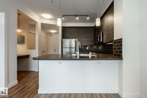 Modern kitchen featuring dark wood-finish cabinetry, stainless steel appliances, and a dark subway tile backsplash - #215 2590 Anderson Wy Sw, Edmonton, AB - Indoor Photo Showing Kitchen With Upgraded Kitchen