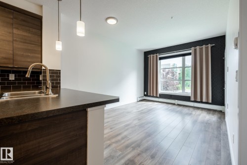 Modern kitchen peninsula featuring a stainless steel sink with a gooseneck faucet, dark countertops, dark wood-finish cabinetry, and a dark subway tile backsplash - #215 2590 Anderson Wy Sw, Edmonton, AB - Indoor Photo Showing Kitchen With Double Sink