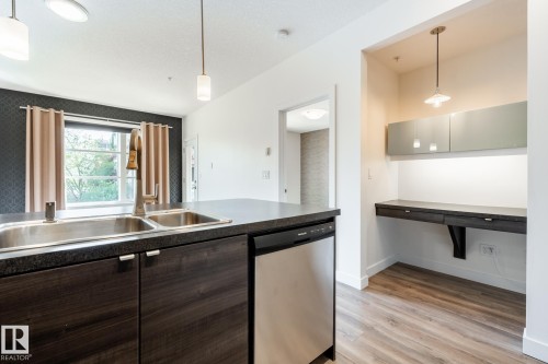 Kitchen island with a double stainless steel sink, dark wood-finish cabinetry, and an integrated dishwasher - #215 2590 Anderson Wy Sw, Edmonton, AB - Indoor Photo Showing Kitchen With Double Sink