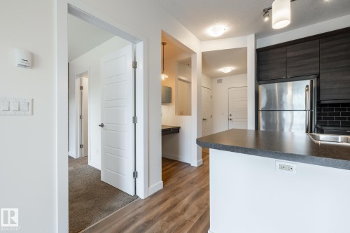 Kitchen island with a dark countertop and integrated sink, stainless steel refrigerator, dark wood-finish cabinetry, black subway tile backsplash, and wood-finish flooring - #215 2590 Anderson Wy Sw, Edmonton, AB - Indoor