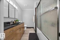 Bathroom featuring a double vanity with a dark countertop and undermount sinks, wood-finish cabinetry, and a shower with sliding textured glass doors - 