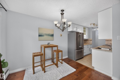 Kitchen and dining area featuring wood-finish flooring, a contemporary chandelier, white cabinetry, stainless steel appliances, and a tiled backsplash - #602 9725 106 St Nw, Edmonton, AB 