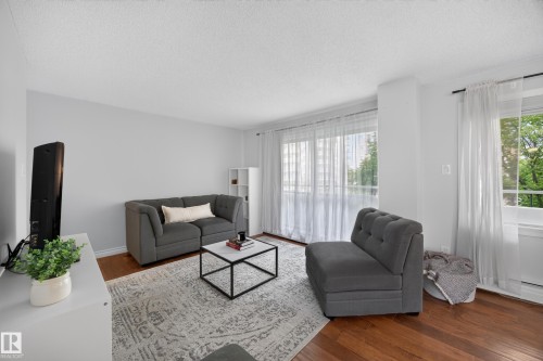 Living area featuring wood-finish flooring, light-colored walls, and large windows with sheer privacy curtains - #602 9725 106 St Nw, Edmonton, AB 