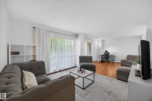 Open-concept living area featuring a large window with sheer white curtains, wood-finish flooring, and a white built-in shelving unit - #602 9725 106 St Nw, Edmonton, AB 