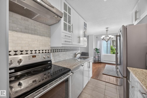 Kitchen featuring a stainless steel electric range, a mosaic tile backsplash with decorative border, white cabinetry, and granite countertops - #602 9725 106 St Nw, Edmonton, AB 