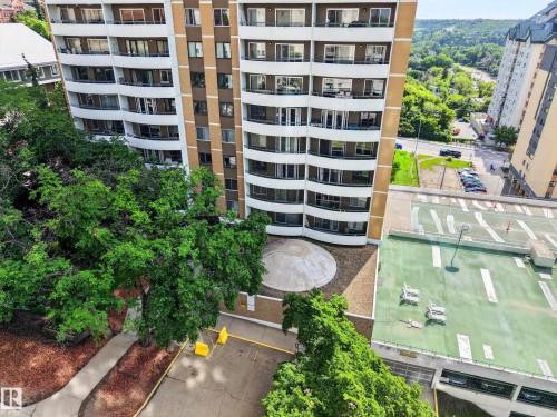 Multi-story residential building featuring private balconies, tan and brown exterior paneling, mature trees, and an adjacent parking structure with a green top surface - #602 9725 106 St Nw, Edmonton, AB 