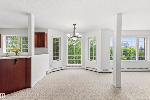 Bright interior space featuring multiple large windows with grid patterns, carpet flooring, and a chandelier light fixture - #201 14 Mission Av, St. Albert, AB - Indoor