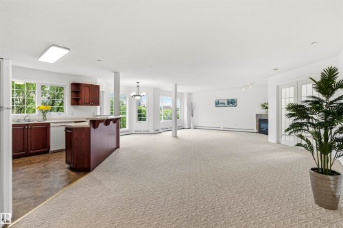 Spacious open-concept living area featuring light-toned patterned carpeting, multiple large windows, a corner fireplace, and double French doors - #201 14 Mission Av, St. Albert, AB - Indoor Photo Showing Other Room