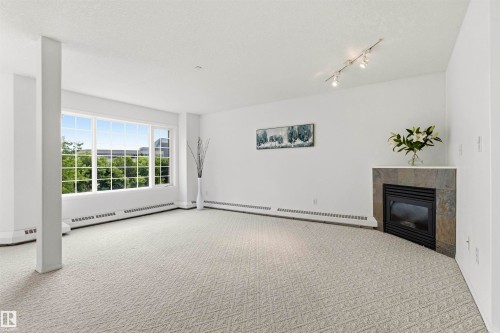 Spacious room featuring broad windows with exterior greenery, wall-to-wall patterned carpeting, a corner fireplace with a stone surround and hearth, track lighting, and baseboard heating - #201 14 Mission Av, St. Albert, AB - Indoor With Fireplace