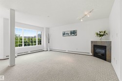 Spacious room featuring broad windows with exterior greenery, wall-to-wall patterned carpeting, a corner fireplace with a stone surround and hearth, track lighting, and baseboard heating - 