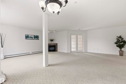 Expansive carpeted room featuring a ceiling-mounted light fixture, French doors with divided lites, a built-in fireplace with a stone surround, and baseboard heating - #201 14 Mission Av, St. Albert, AB - Indoor Photo Showing Other Room