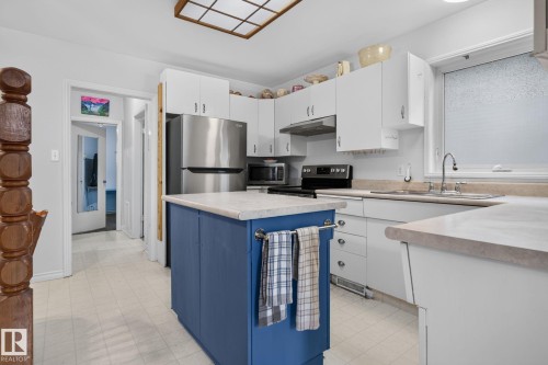 Kitchen featuring a blue island with storage, white cabinetry, integrated stainless steel appliances, a double basin sink, and a multi-paneled ceiling light fixture - 12703 122 Street Nw, Edmonton, AB 