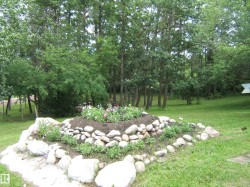 Expansive green space featuring a tiered rock garden with plantings, surrounded by mature trees and verdant foliage - 