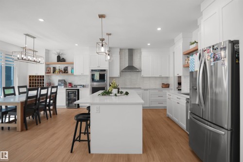 Bright open-concept kitchen featuring white shaker cabinetry, stainless steel appliances, a center island with countertop seating, wood-finish flooring, and a dining area with a built-in wine rack - 5802 Soleil Boulevard, Beaumont, AB 