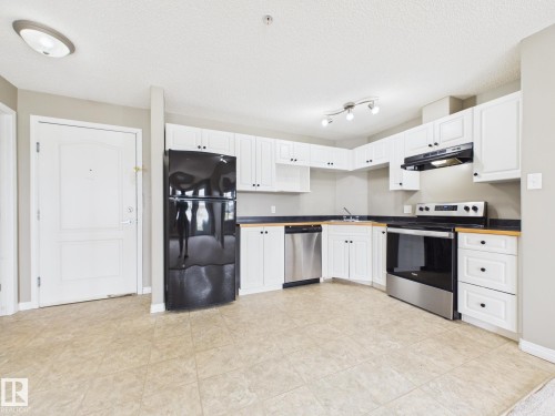 Kitchen featuring light-toned flooring, white cabinetry, dark countertops, a stainless steel dishwasher, and a black refrigerator - #309 5350 199 St Nw, Edmonton, AB 