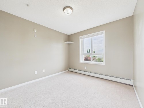 Neutral-toned room featuring a large window, light-colored carpet, and wall-mounted baseboard heating - #309 5350 199 St Nw, Edmonton, AB 