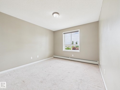 Neutral-toned interior featuring wall-to-wall carpeting, a large window with white trim, and baseboard heating - #309 5350 199 St Nw, Edmonton, AB 