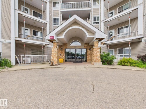 Building exterior featuring a covered entrance with stone-clad pillars, glass double doors, and multiple balconies with white railings - #309 5350 199 St Nw, Edmonton, AB 