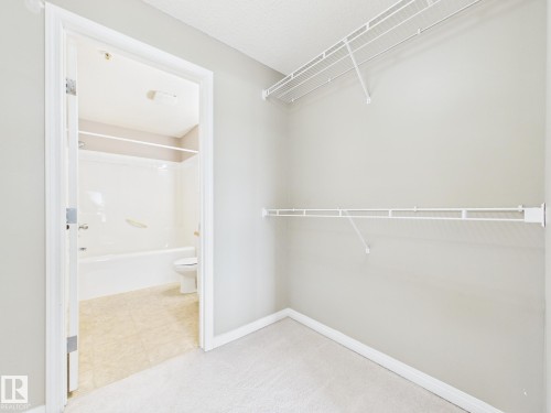 Carpeted closet with wire shelving and a doorway opening to a bathroom featuring a white bathtub, toilet, and tile flooring - #309 5350 199 St Nw, Edmonton, AB 