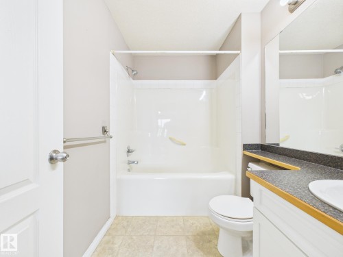 Full bathroom featuring a white tub and shower combination, a white toilet, a vanity with a dark countertop and integrated sink, and light-toned floor tiling - #309 5350 199 St Nw, Edmonton, AB 