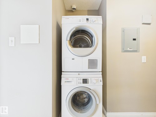 Integrated laundry area featuring a stacked washer and dryer, utility panels, and light-toned wall finishes - #309 5350 199 St Nw, Edmonton, AB 
