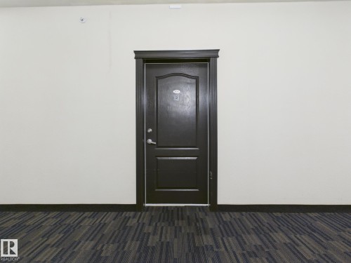 Richly toned door with decorative paneling and polished chrome hardware, framed by dark trim, set against light neutral walls and patterned carpet flooring - #309 5350 199 St Nw, Edmonton, AB 