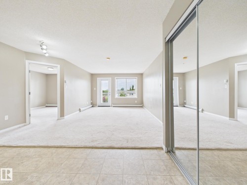 Spacious interior room featuring light-colored carpeting, a balcony door, and an expansive sliding mirrored closet - #309 5350 199 St Nw, Edmonton, AB 