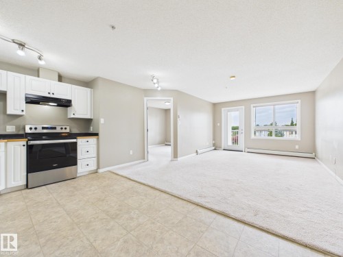 Kitchen and living area featuring white cabinetry with black hardware, a stainless steel and black range, black countertops, and track lighting - #309 5350 199 St Nw, Edmonton, AB 