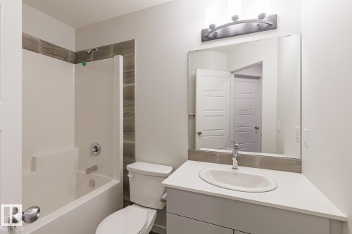 Bathroom featuring a white bathtub/shower combination with a textured tile surround, a single-sink vanity with a white countertop, and a large wall-mounted mirror with an overhead light fixture - 3619 Triomphe Boulevard, Beaumont, AB - Indoor Photo Showing Bathroom