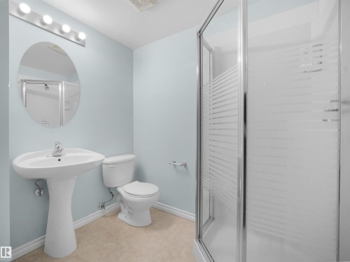 Full bathroom featuring a pedestal sink, a toilet, an oval mirror with vanity lighting, and a glass-enclosed shower with frosted accents - #207 10606 102 Av Nw, Edmonton, AB 