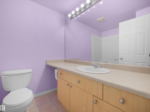 Bathroom featuring a light wood-finish vanity with a built-in sink, an expansive wall mirror with overhead lighting, a white toilet, and tiled flooring - #207 10606 102 Av Nw, Edmonton, AB 