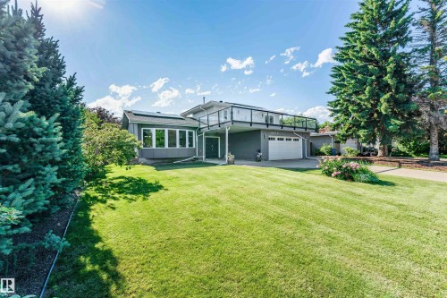 Modern gray exterior featuring a sunroom with multiple windows, an upper-level deck with glass railing, an integrated garage, and a lush green lawn - 5212 142 Street Nw, Edmonton, AB - Outdoor